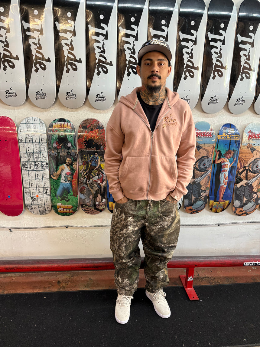 Person wearing a pink hoodie and camouflage pants standing in front of a wall with skateboard decks and 'Trade' branding.