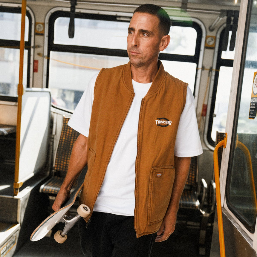 Man in a brown vest and white shirt holding a skateboard inside a bus.