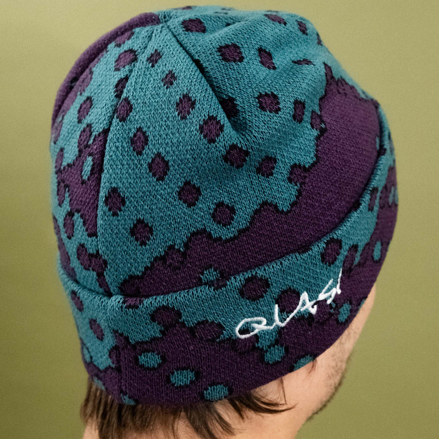 Teal and purple patterned beanie with a brand logo on a green background