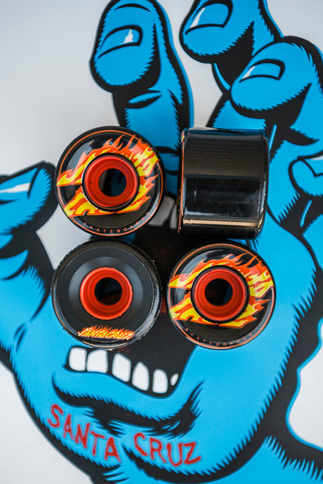 Four skateboard wheels with flame design on a blue hand graphic background, featuring the Santa Cruz brand.