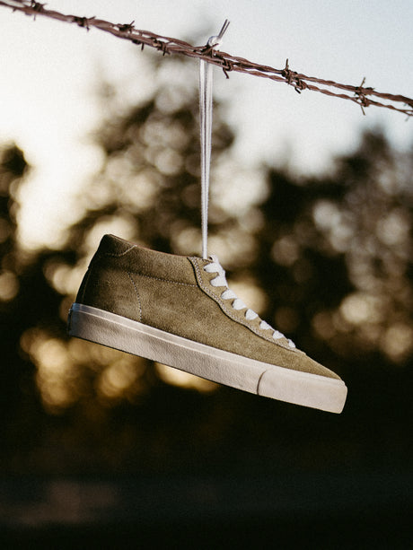 Brown suede sneaker with white sole hanging on barbed wire against a blurred natural background